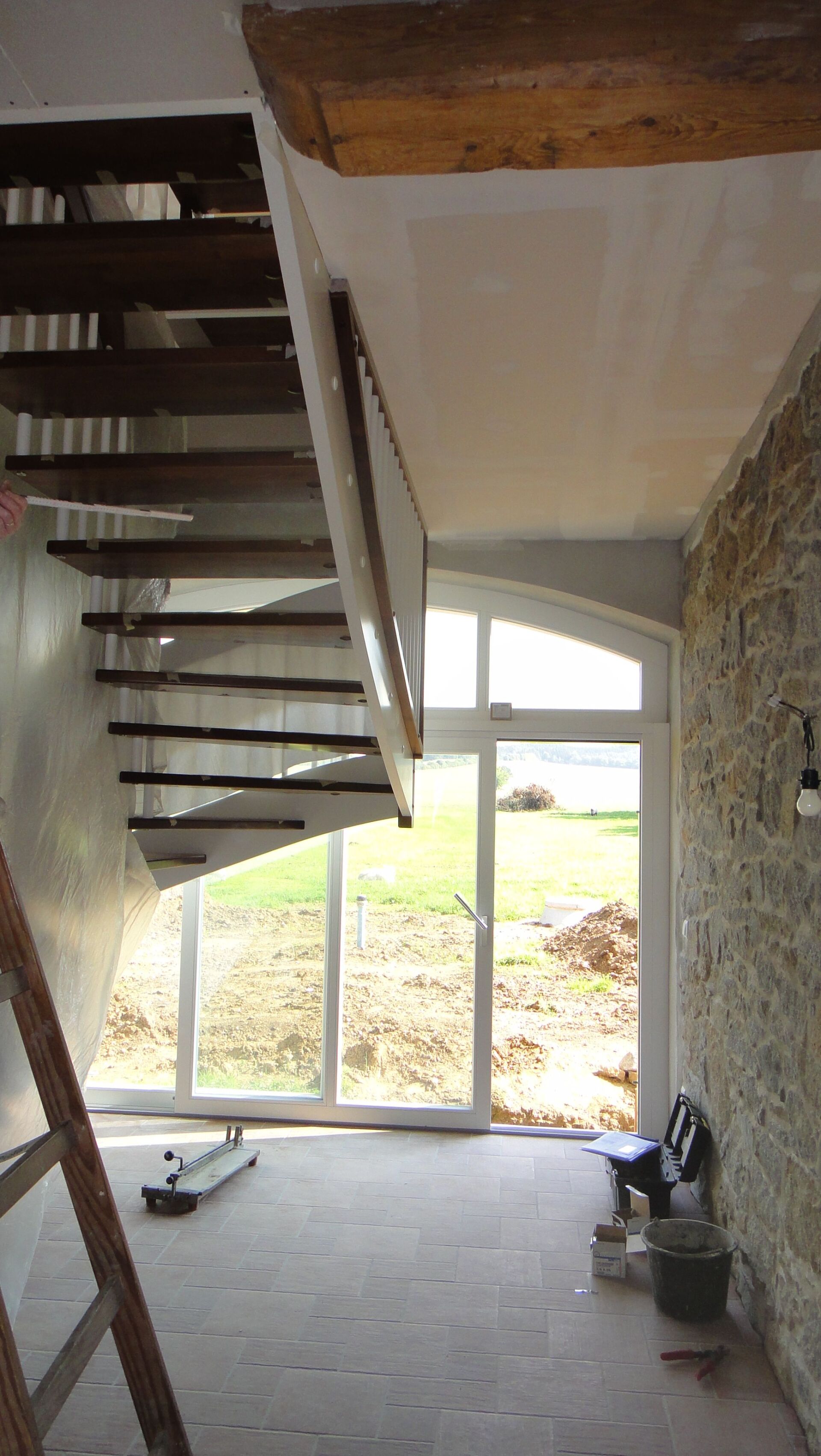 Innenansicht eines Treppenhauses mit Tür und Fenstern. Treppe mit Holzstufen und Backsteinwand. Helles Tageslicht fällt durch die Fenster.