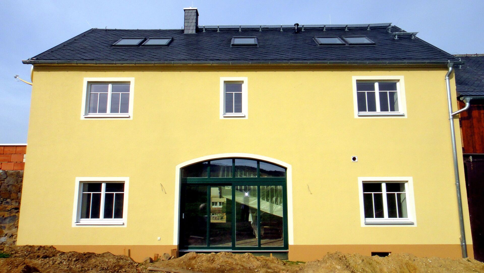 Gelbes zweistöckiges Gebäude mit dunklem Dach, gläsernem Bogeneingang und weiß gerahmten Fenstern vor blauem Himmel.