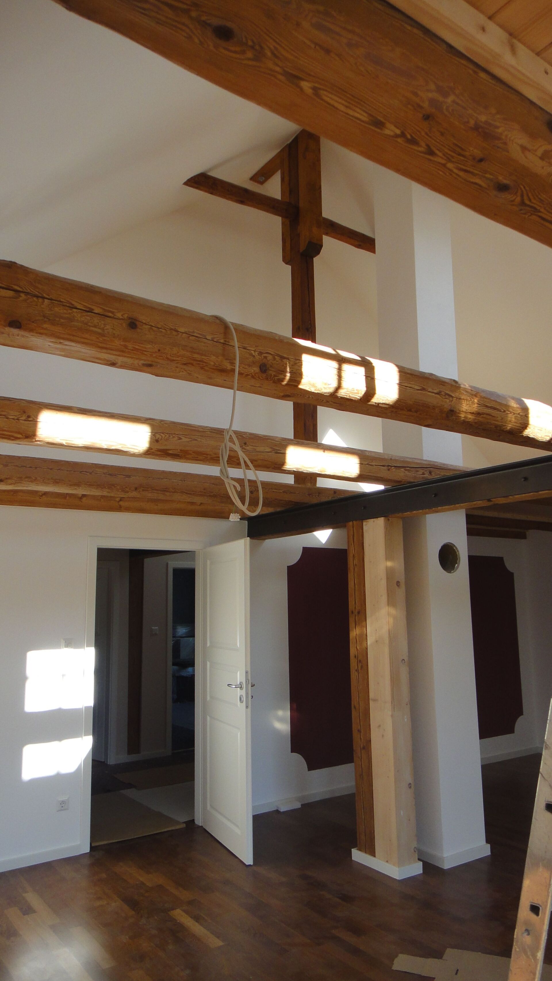 Innenansicht eines Zimmers mit freiliegenden Holzbalken, weißen Wänden und einer Tür, die in einen anderen Raum führt.