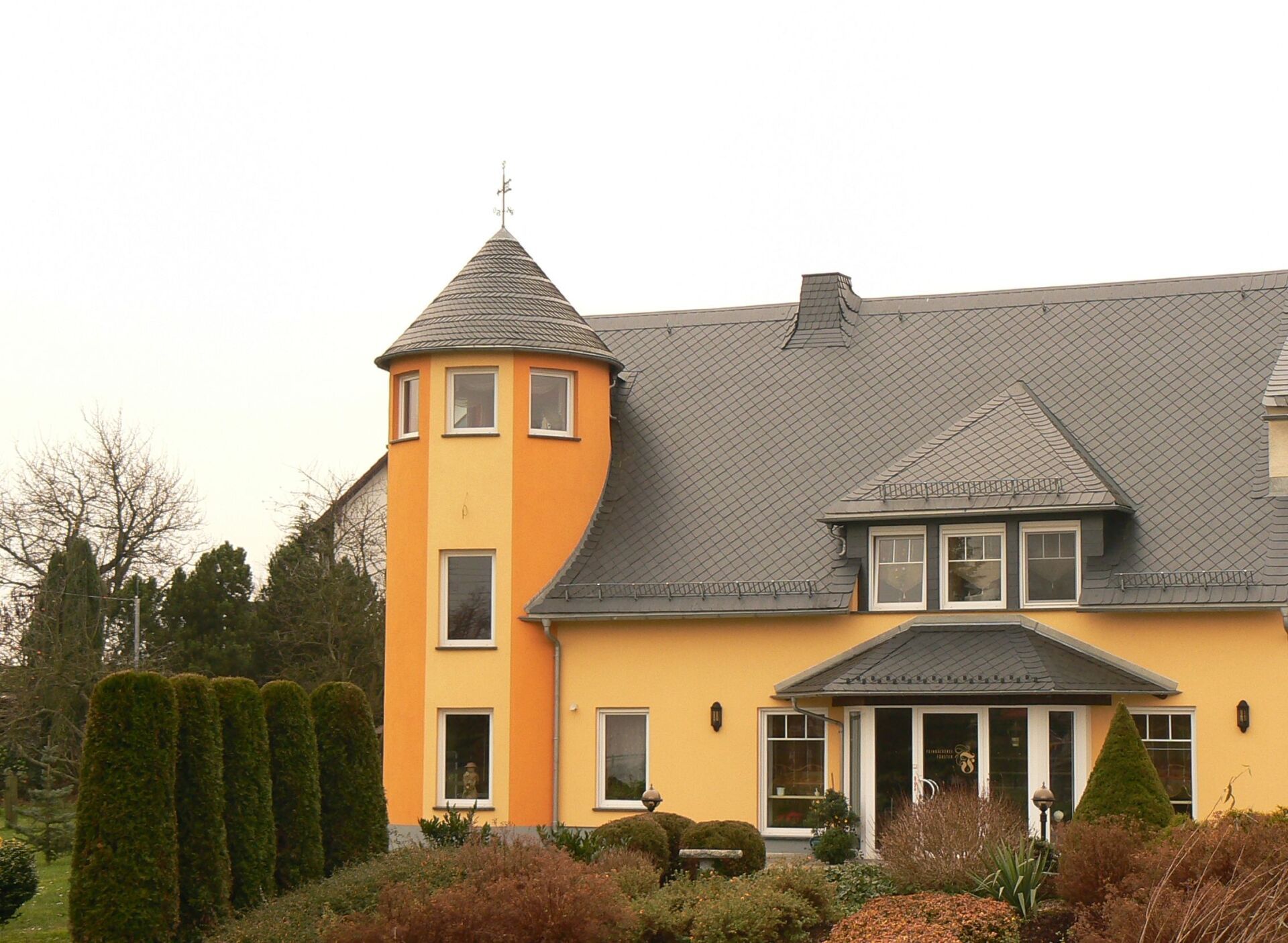 Gelbes Haus mit Turm, graues Dach, umgeben von grünen Sträuchern, unter einem bedeckten Himmel.