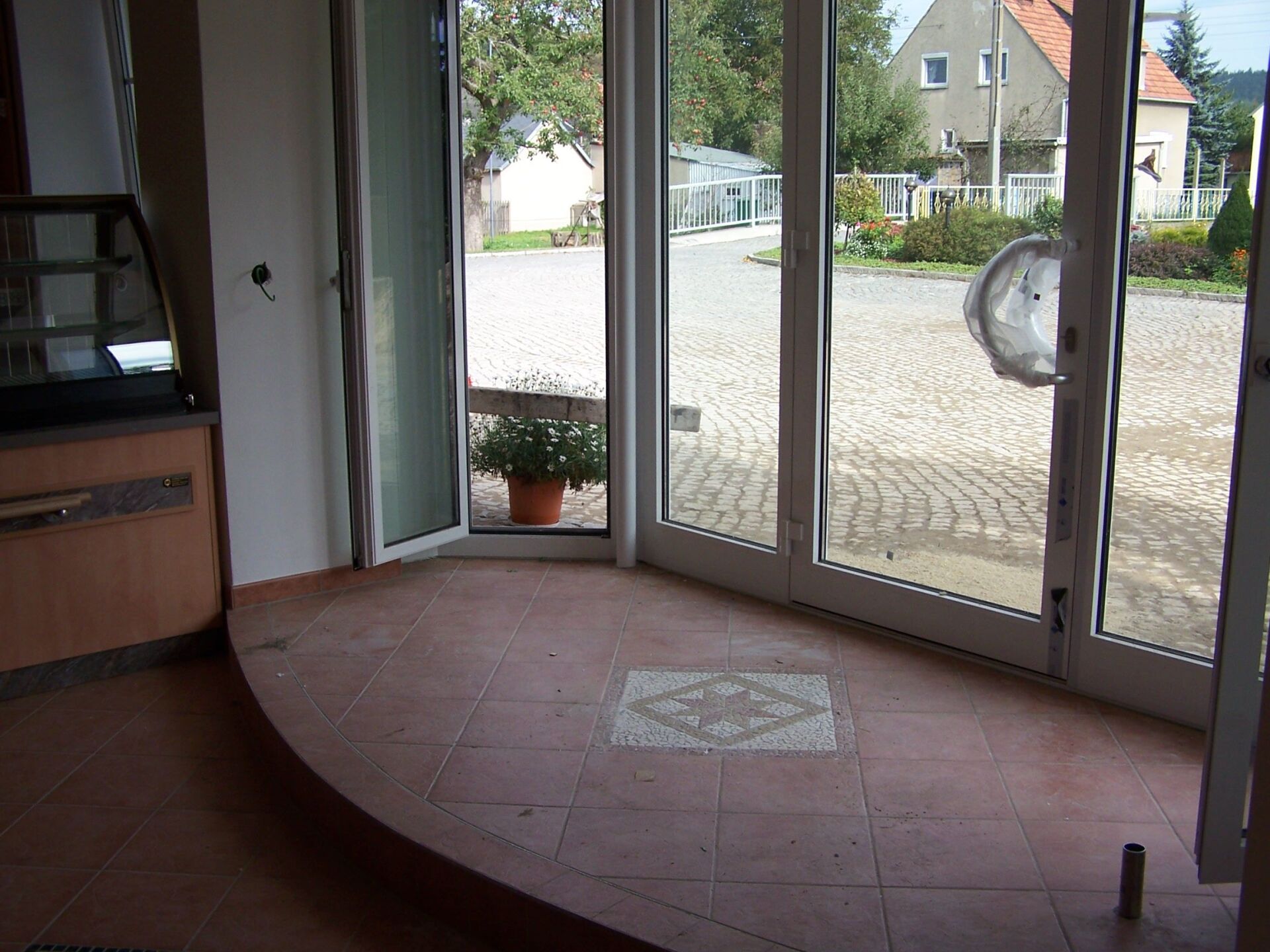 Blick vom Gebäudeinneren nach draußen. Glastüren, Fliesenboden, Topfpflanze sichtbar.