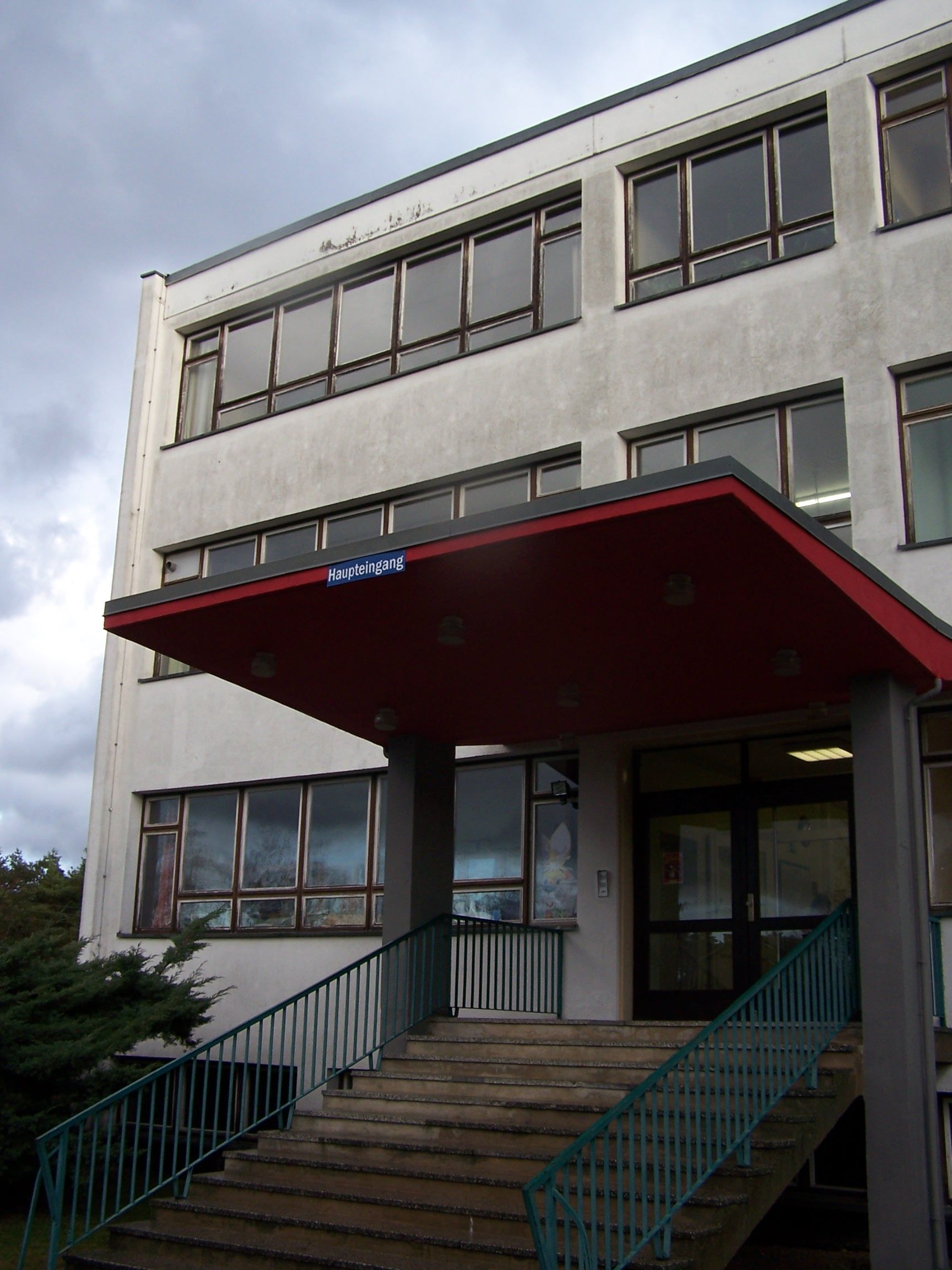 Eingang zum Schulgebäude mit roter Markise, Treppe und Fenstern. Bewölkter Himmel.