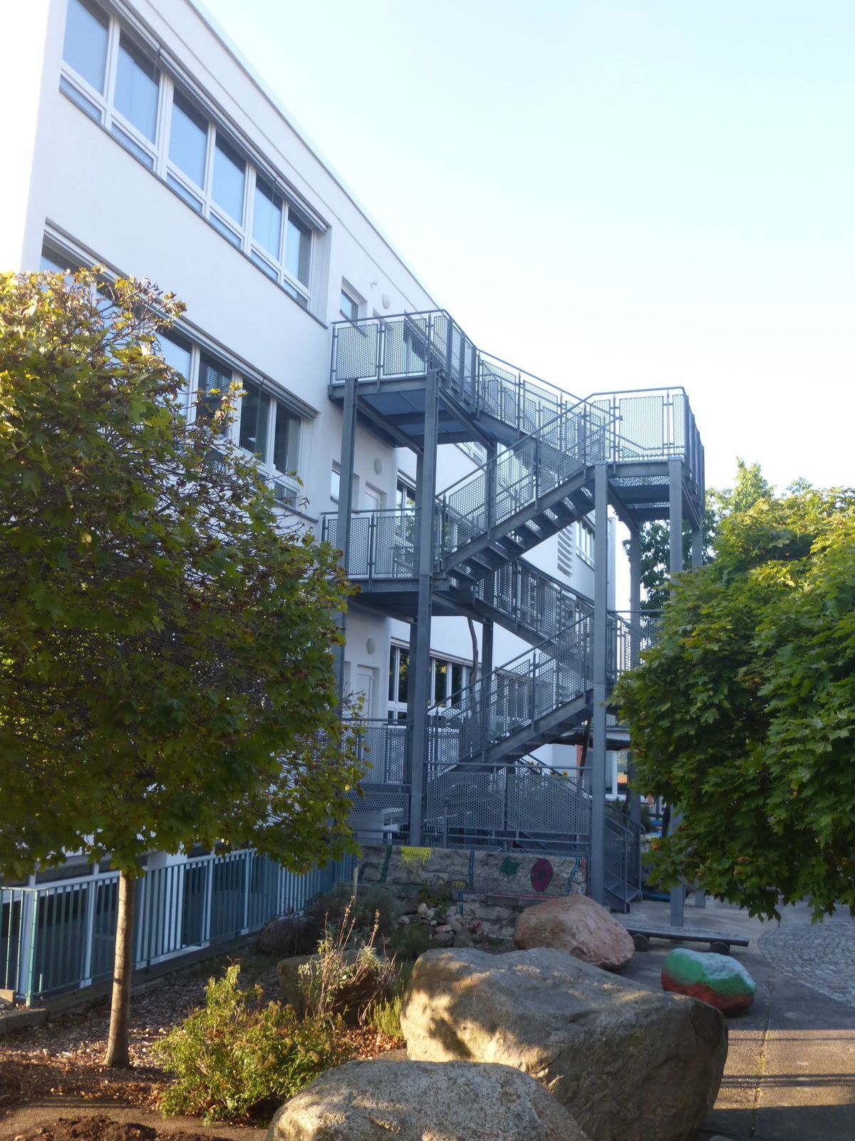 Außenansicht eines Gebäudes mit einer metallenen Feuertreppe. Im Vordergrund sind Bäume und große Felsen zu sehen.