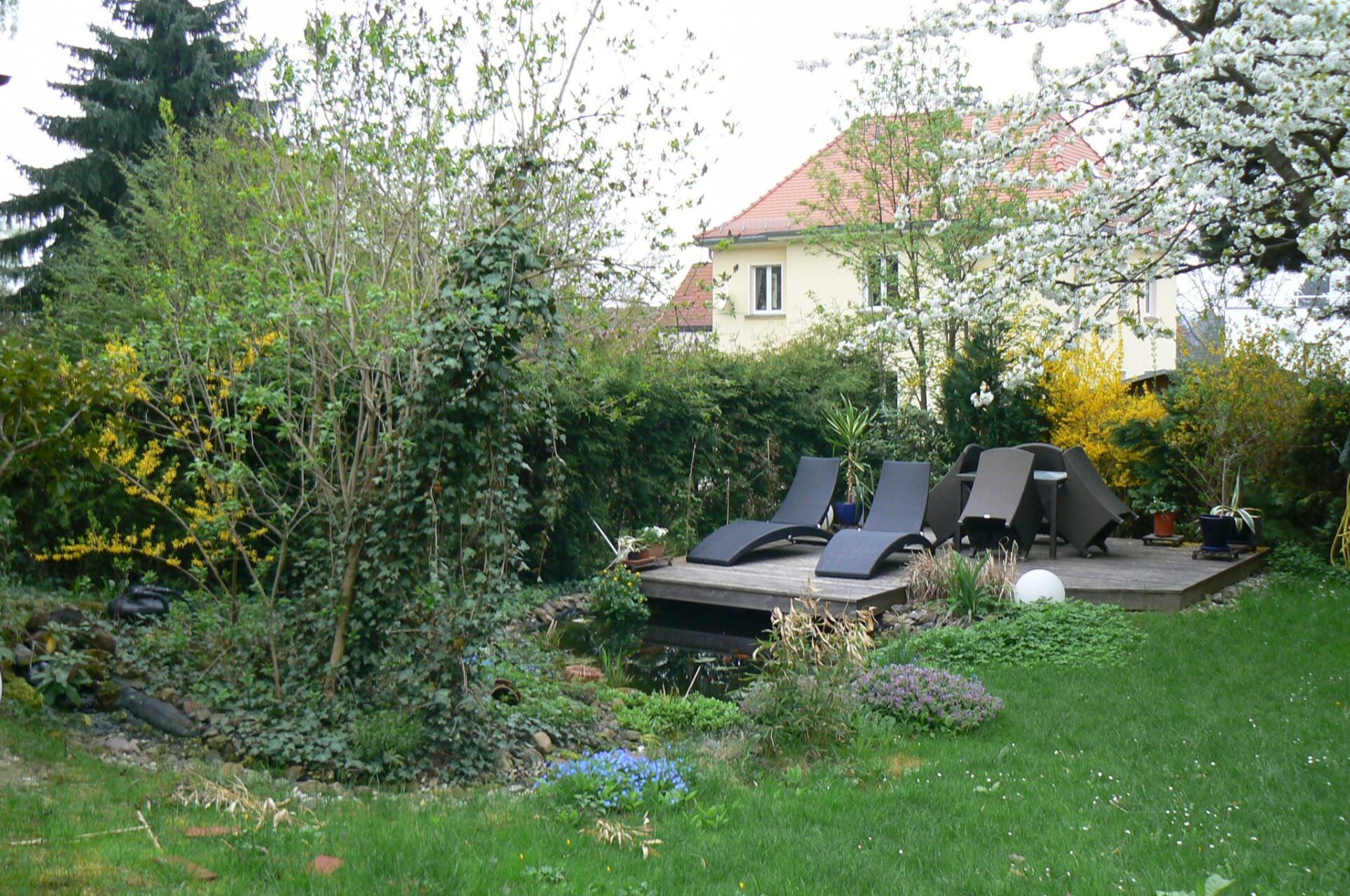 Liegestühle auf einer Holzterrasse an einem kleinen Teich in einem üppigen Garten, im Hintergrund ein Haus.