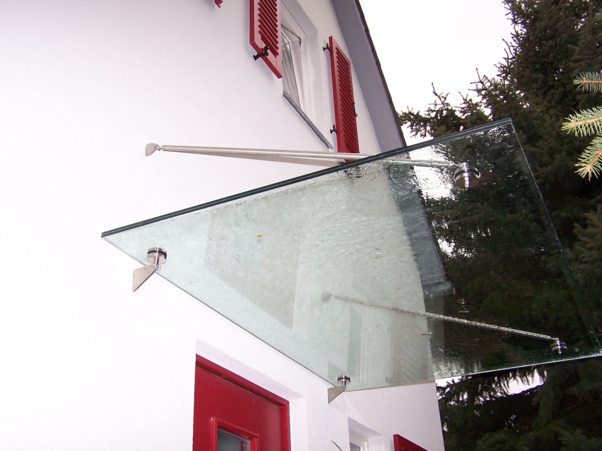 Ein Glasvordach über einer roten Tür, das von Metallarmen getragen wird, die an einem weißen Gebäude befestigt sind.