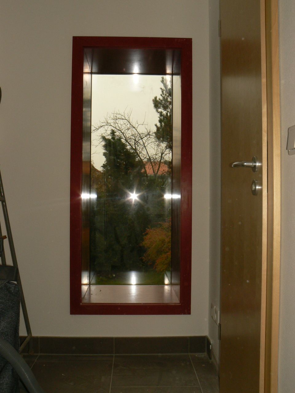 Rechteckiges Fenster mit dunkelrotem Rahmen, Blick auf Bäume und einen bewölkten Himmel; daneben eine helle Holztür.