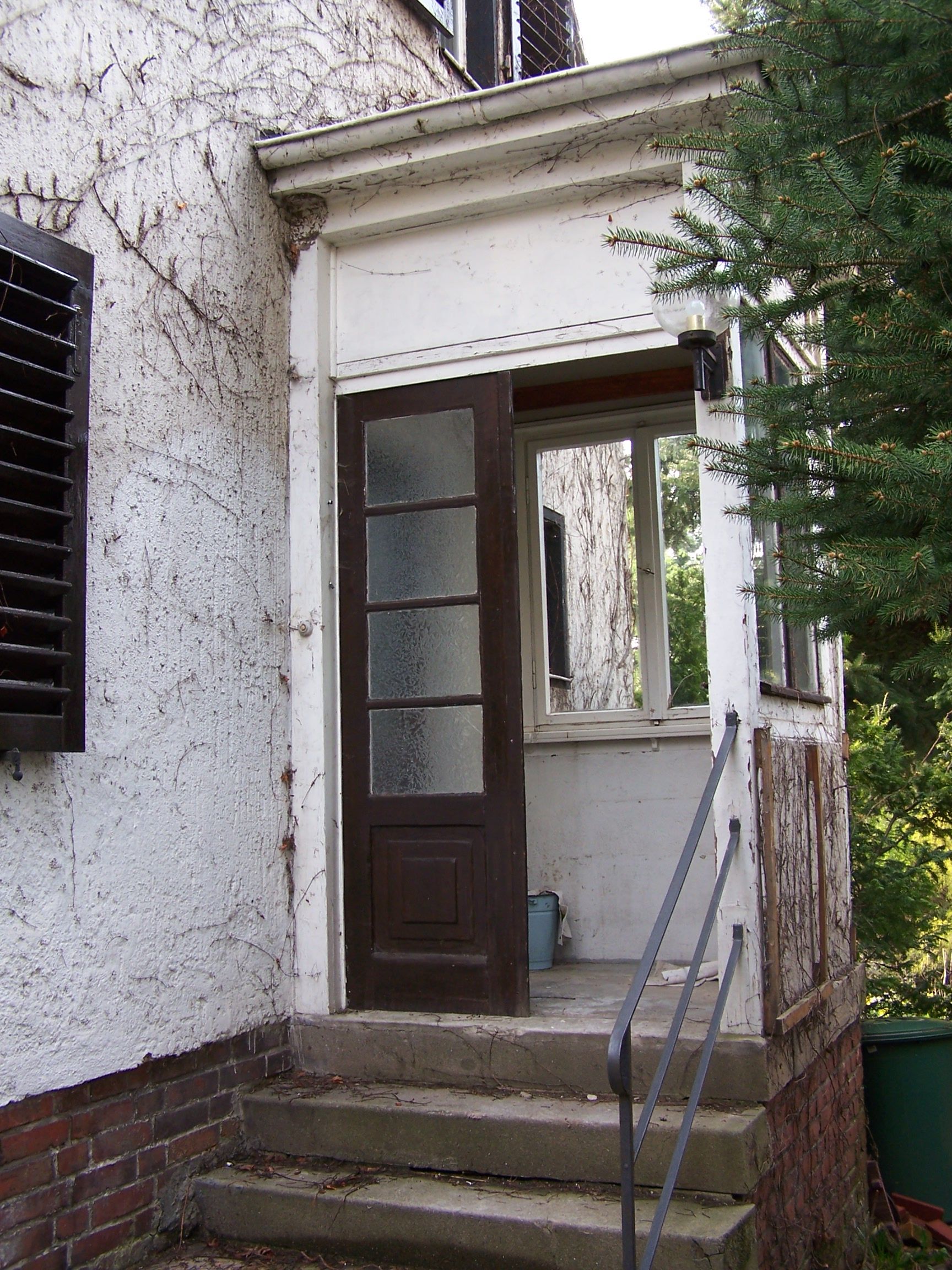 Braune Tür mit Glasscheiben, drei Stufen hinauf. Veranda, weiße Fassade, Efeu, Fensterläden, Geländer.