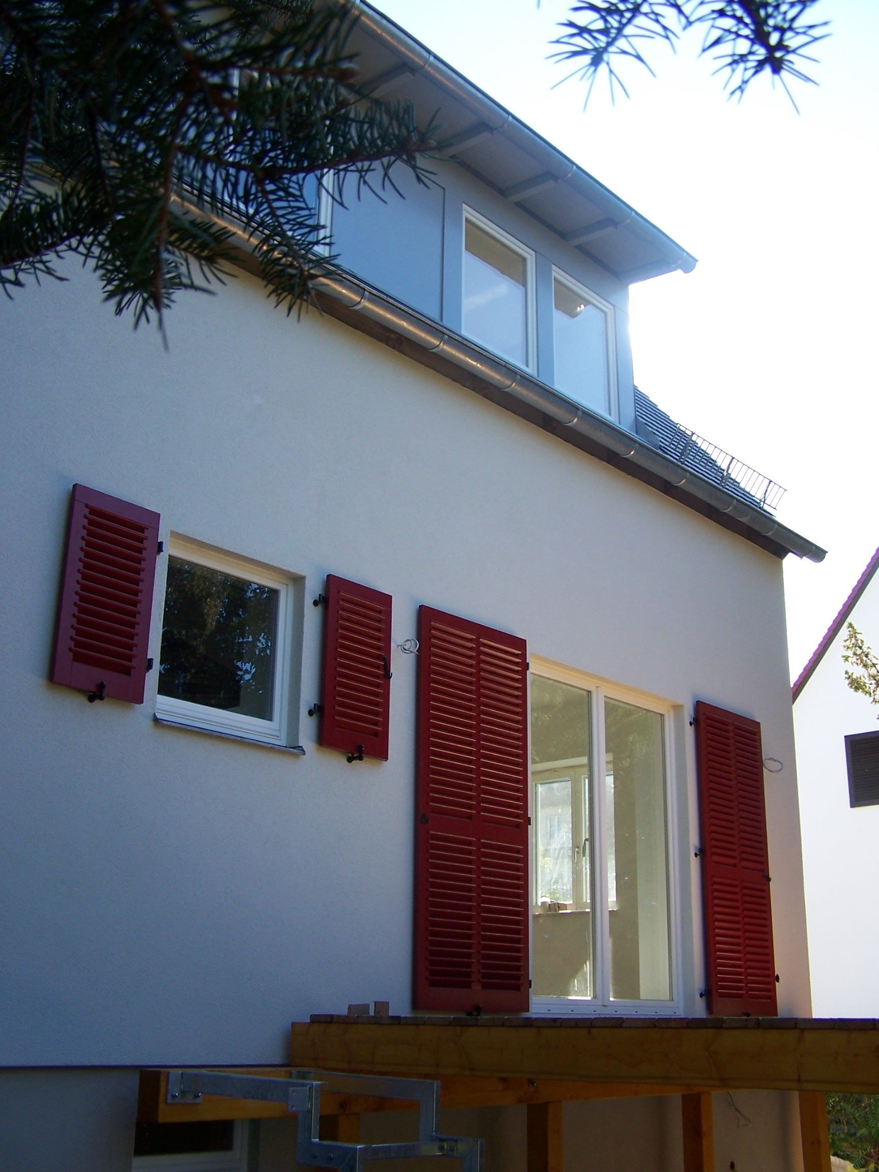 Weißes Haus mit roten Fensterläden und Dachgaube. Holzterrasse davor.