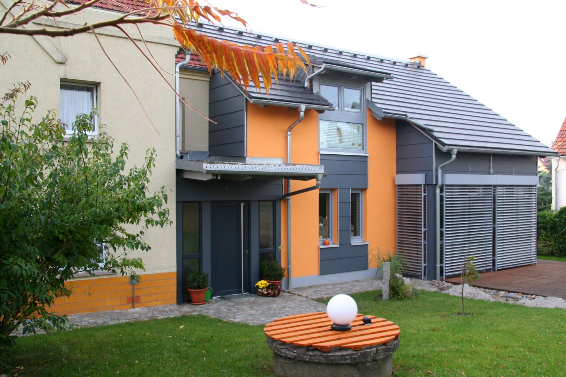 Modernes Haus mit orange-grauer Fassade, überdachtem Eingang und Garage. Grüner Rasen mit Brunnen.