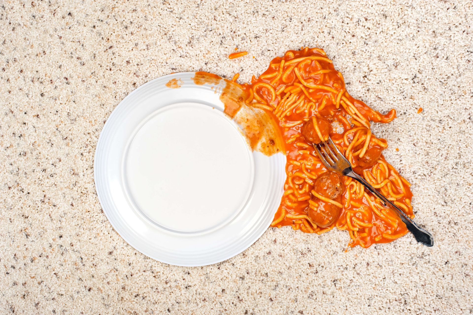 Spilled spaghetti and meatballs with sauce on a light countertop next to an empty white plate and fork.