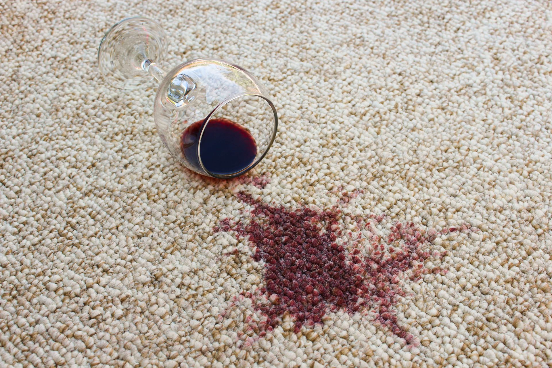 Red wine spilled on a beige carpet, with the overturned glass nearby.