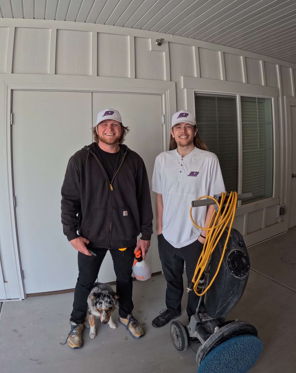 Two men in caps, next to cleaning equipment, a small dog at their feet, in a doorway. Deep Clean Dynamics Carpet Cleaning Company
