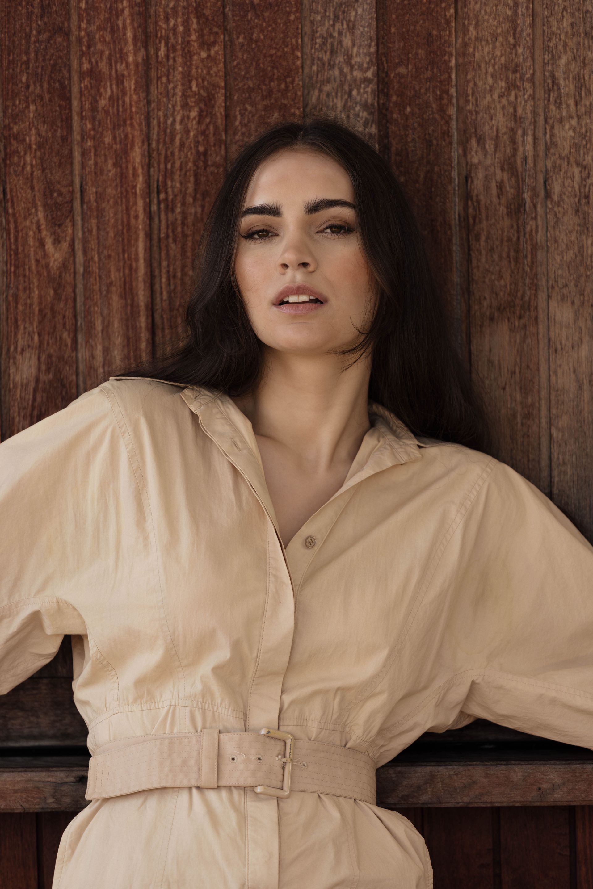 A woman in a beige dress is leaning against a wooden wall.