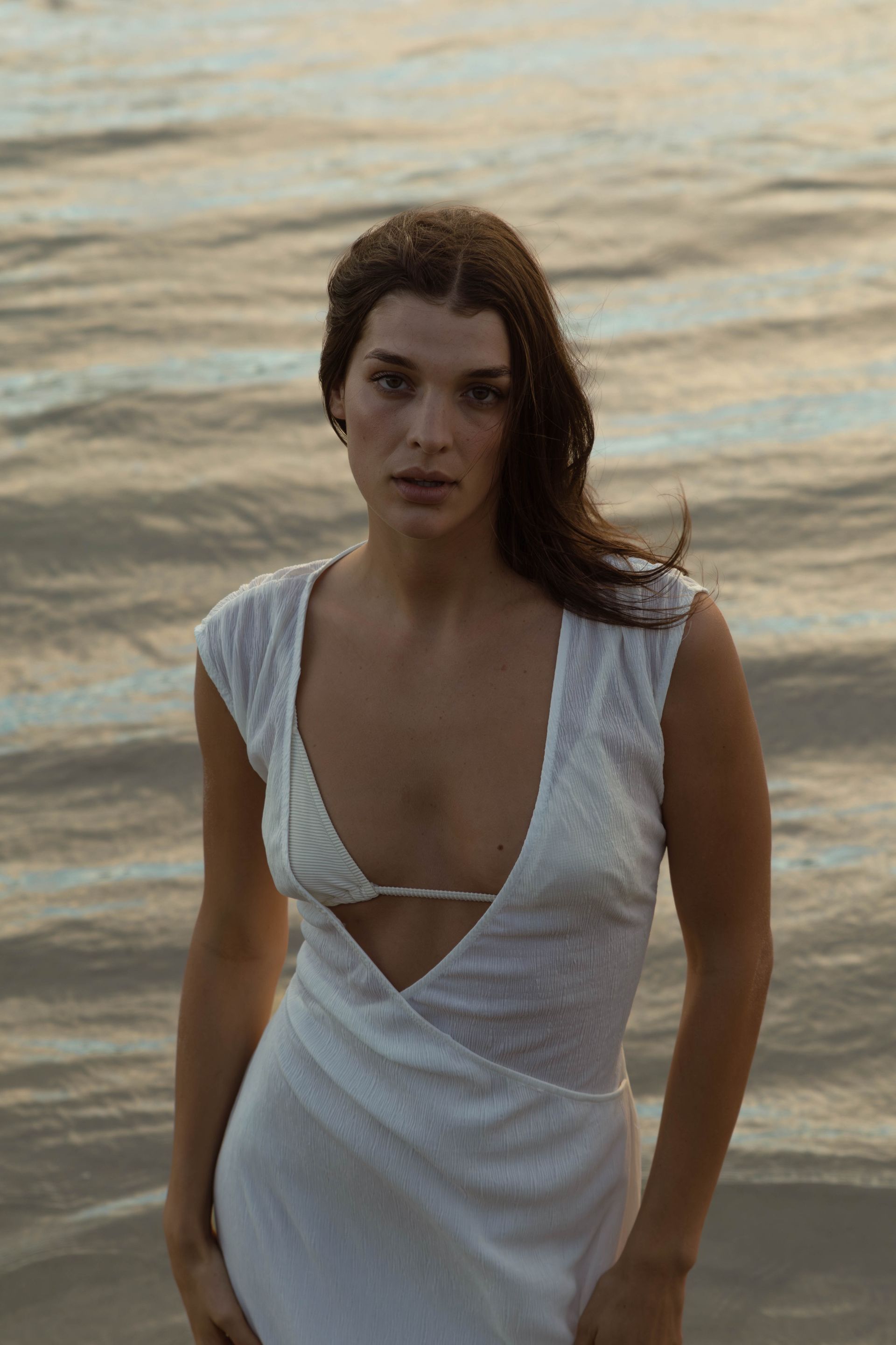 A woman in a white dress is standing on a beach near the water.