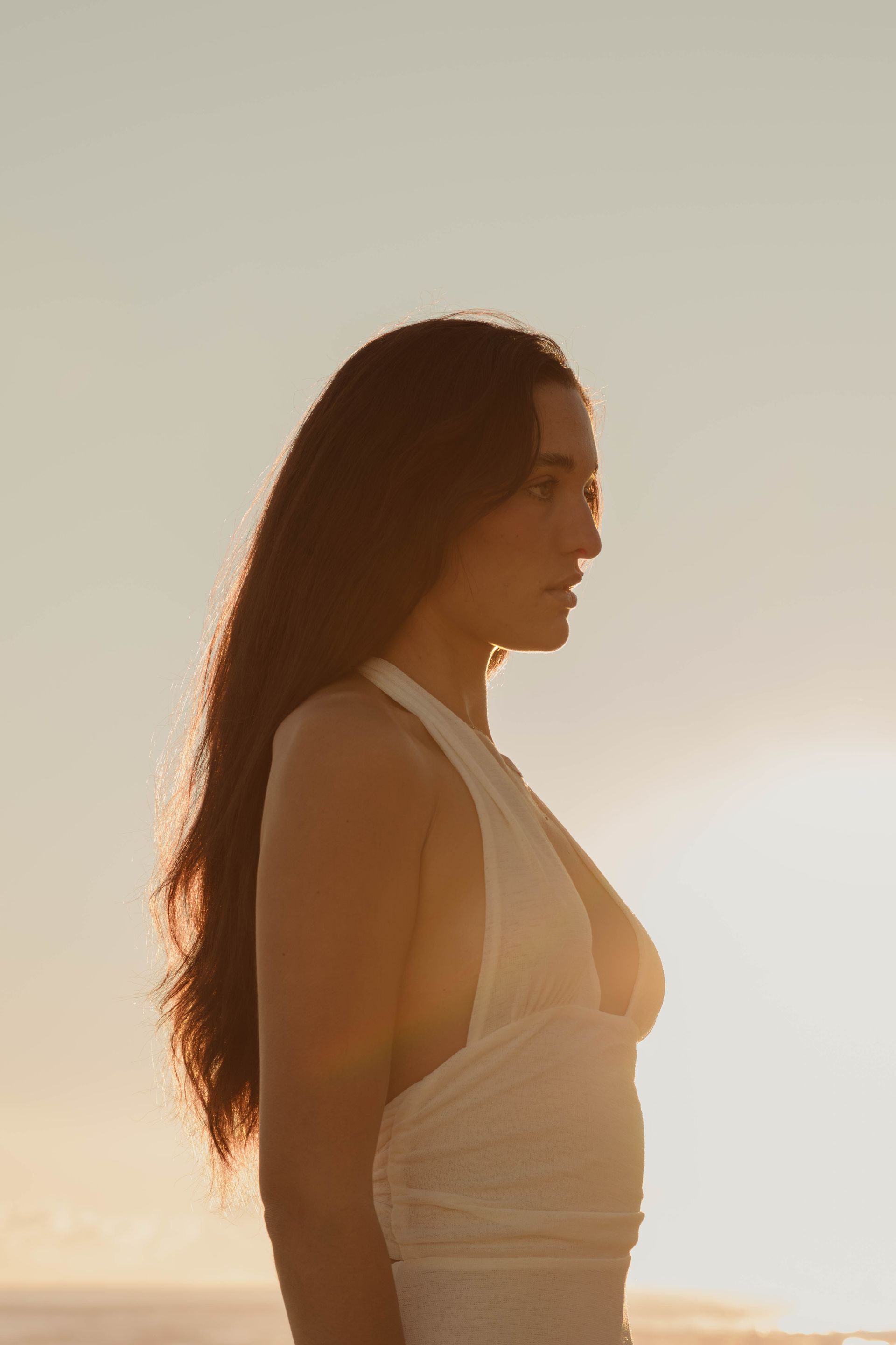 A woman in a white dress is standing on a beach at sunset.