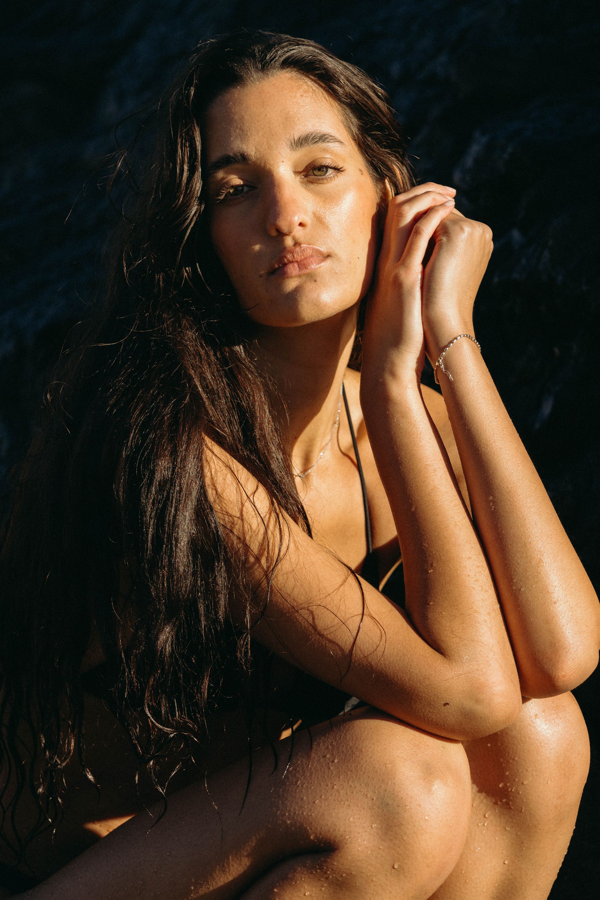 A woman with long hair is sitting on the beach with her hands on her face.