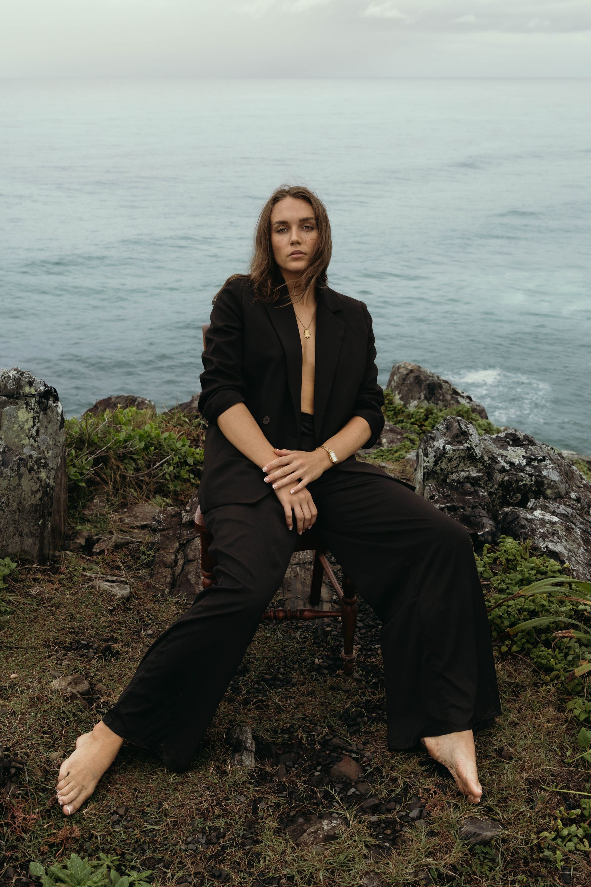 A woman in a black suit is sitting on a rock near the ocean.