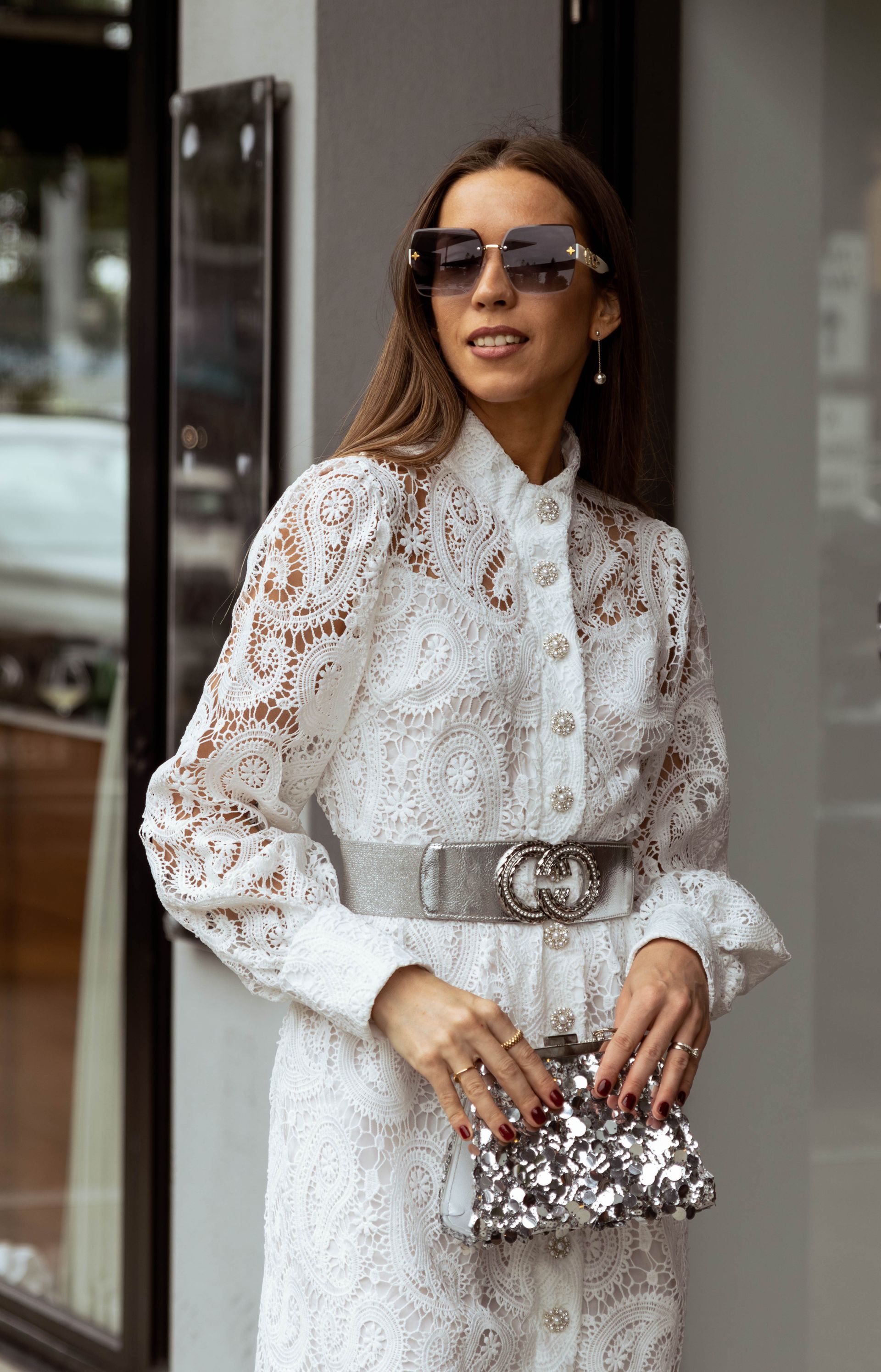 A woman wearing a white lace dress and sunglasses is standing in front of a building.