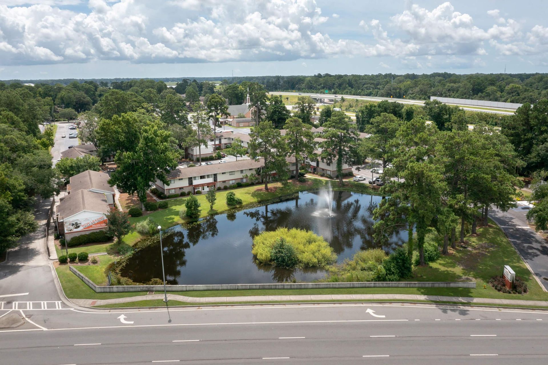 Aerial view of a residential area with a pond, fountain, trees, and buildings, under a cloudy sky.