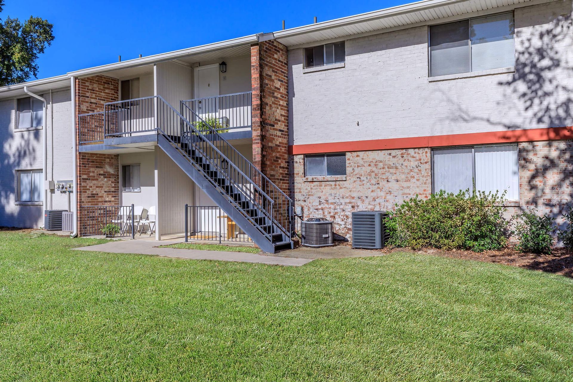 Two-story apartment building with brick and white stucco exterior. Metal staircase, green lawn, and clear blue sky.