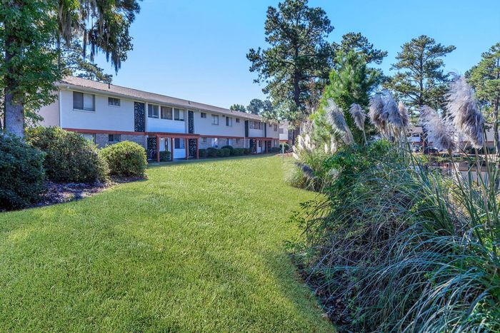 Apartment building with light-colored walls and red trim, set amidst a lush green lawn and tall trees.