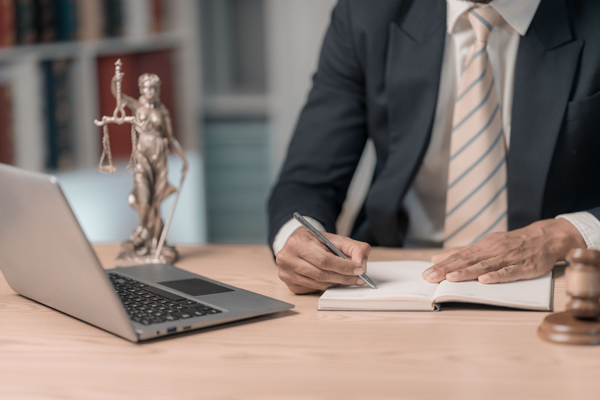 Person writing at desk with laptop, gavel, and Lady Justice statue in legal office. Person writing at desk with laptop, gavel, and Lady Justice statue in legal office.