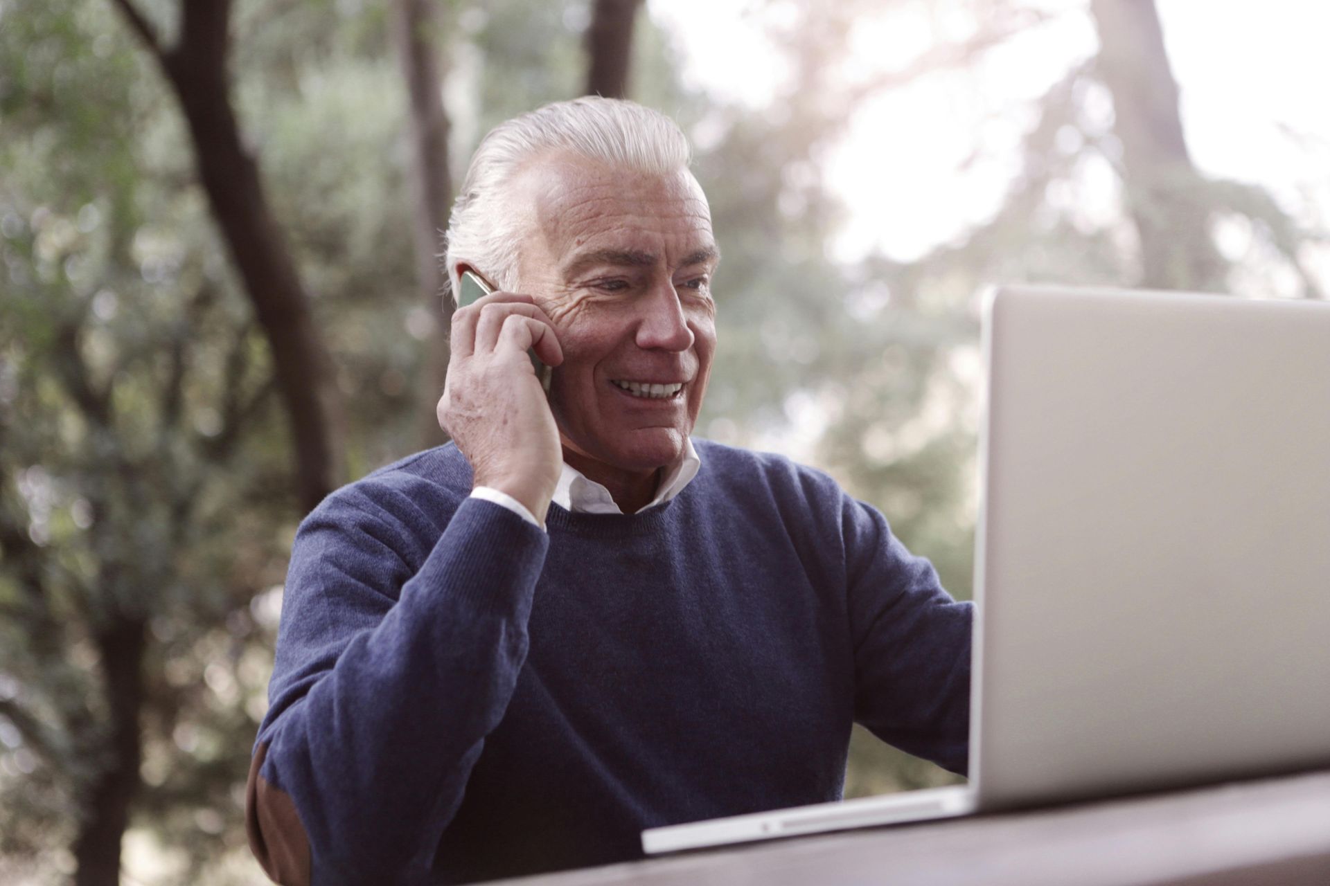 Smiling person talking on a phone while using a laptop outdoors
