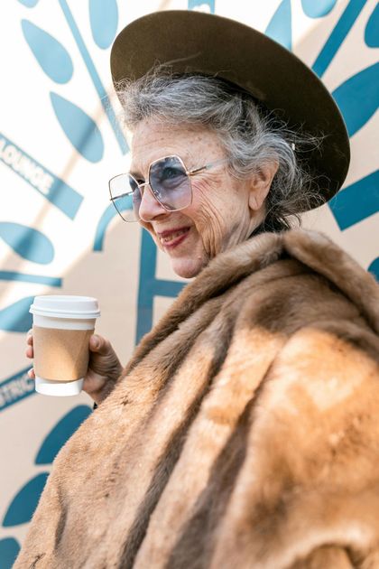Woman in a fur coat and wide-brim hat holding a coffee cup, smiling against a blue mural backdrop