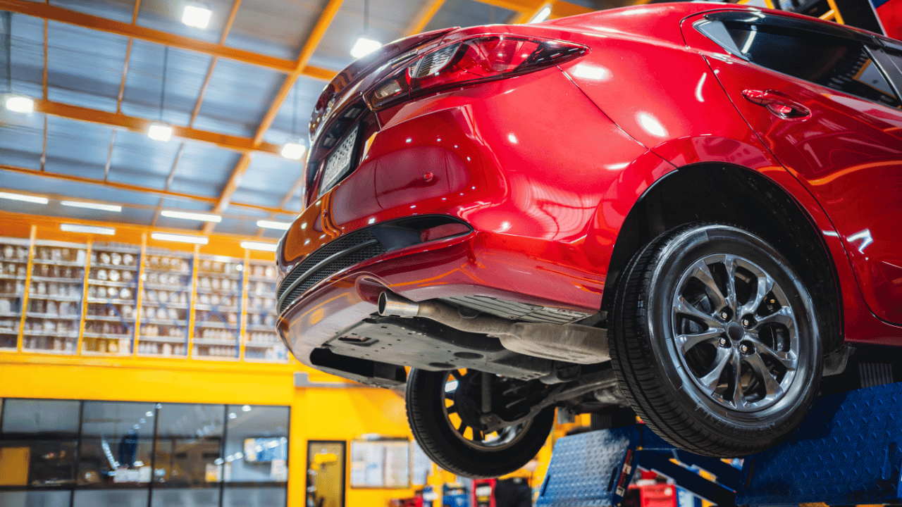 Red car on a lift in a repair shop, yellow walls, tires visible, undercarriage exposed.