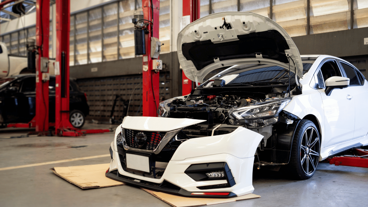 White car with open hood in a repair shop, bumper removed, red lifts in background.