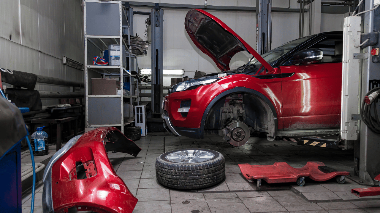 Red car in a repair shop with hood open, wheel off, and parts scattered.