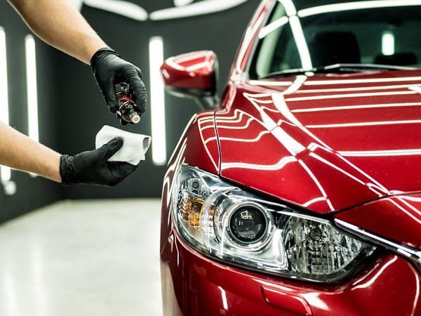 Hands in black gloves applying ceramic coating to a red car's headlight in a well-lit shop.