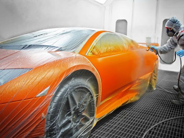 Orange sports car being painted in a paint booth. A person in protective gear sprays the vehicle.