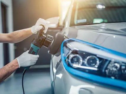 Hands polishing a silver car headlight with a power buffer, indoors.