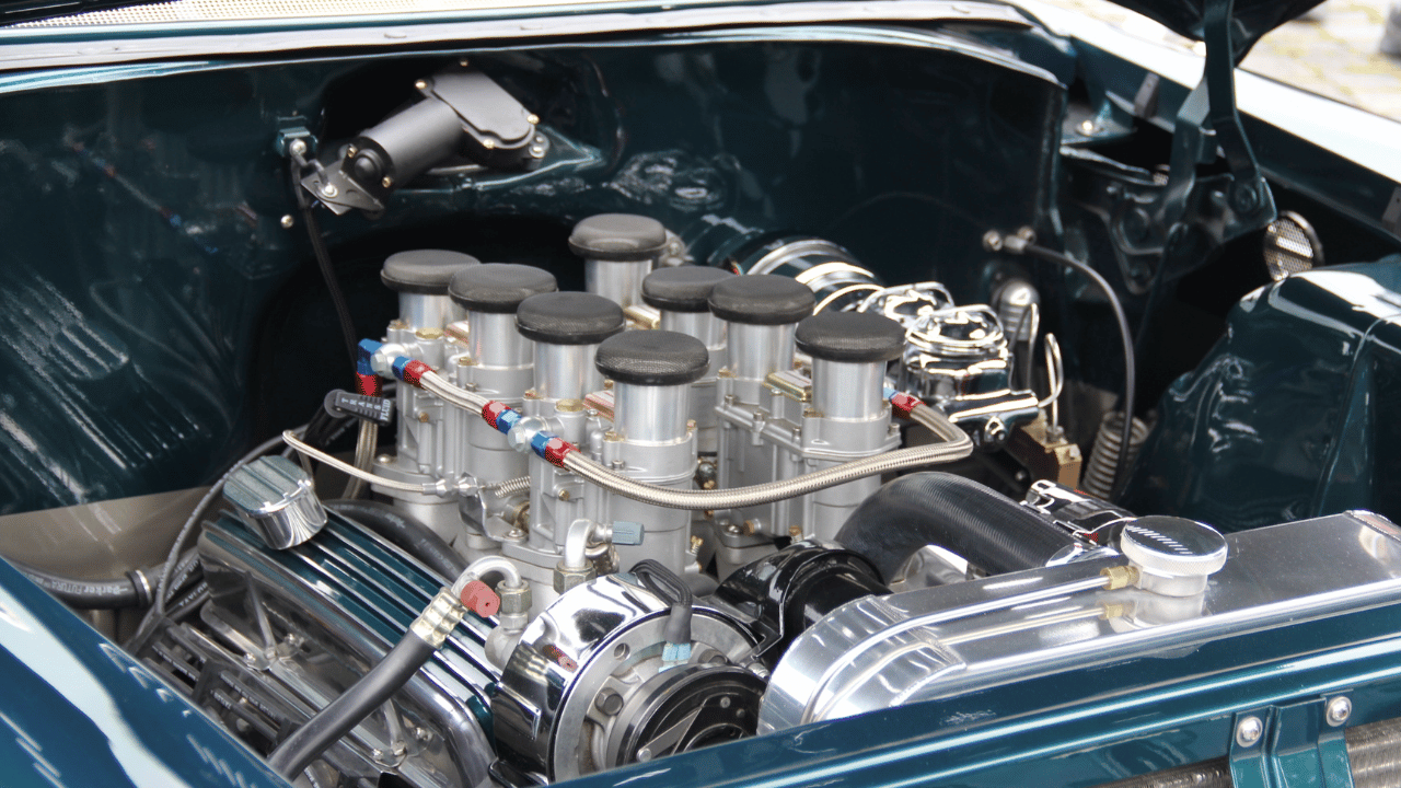 Close-up of a classic car engine with polished chrome and multiple carburetors in a dark teal engine bay.