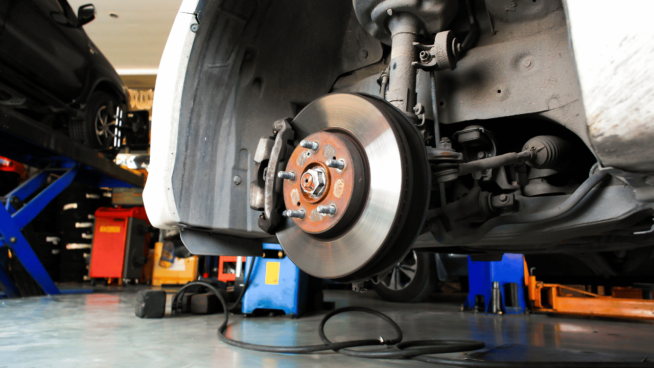 Car's front wheel with brake rotor and caliper visible, raised on a jack in an auto repair shop.
