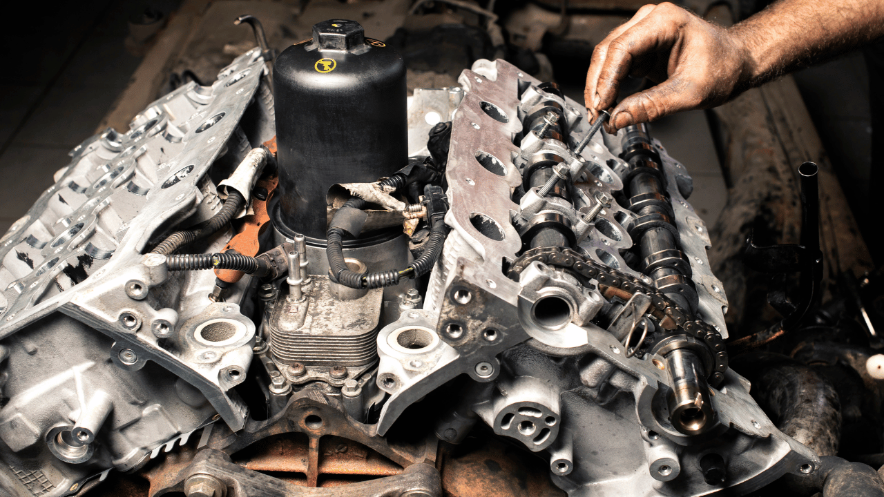 Mechanic's hand working on a disassembled car engine, with visible gears, chains, and components.