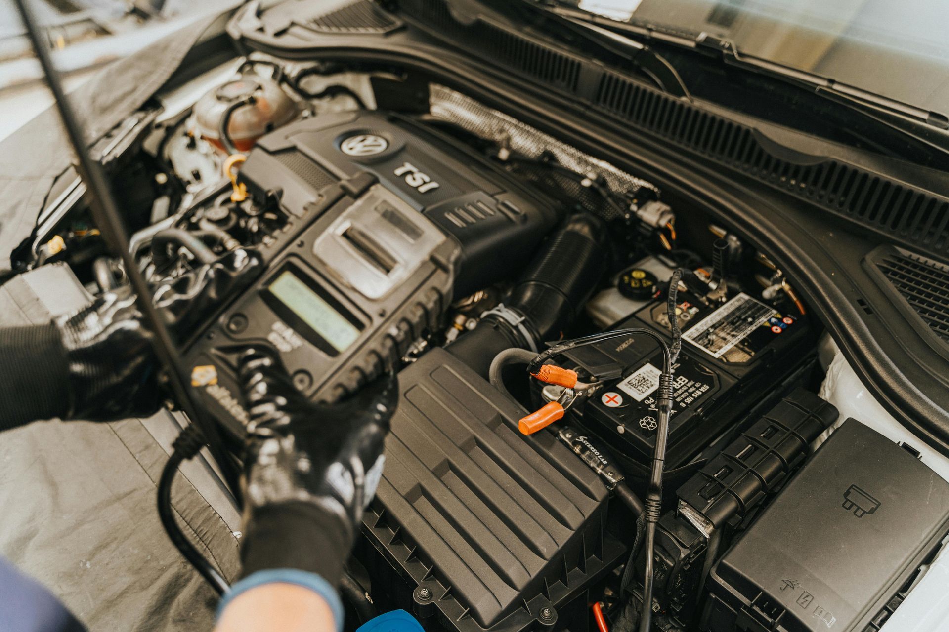 Mechanic using diagnostic equipment on a car engine. Gloves, battery, and engine details visible.