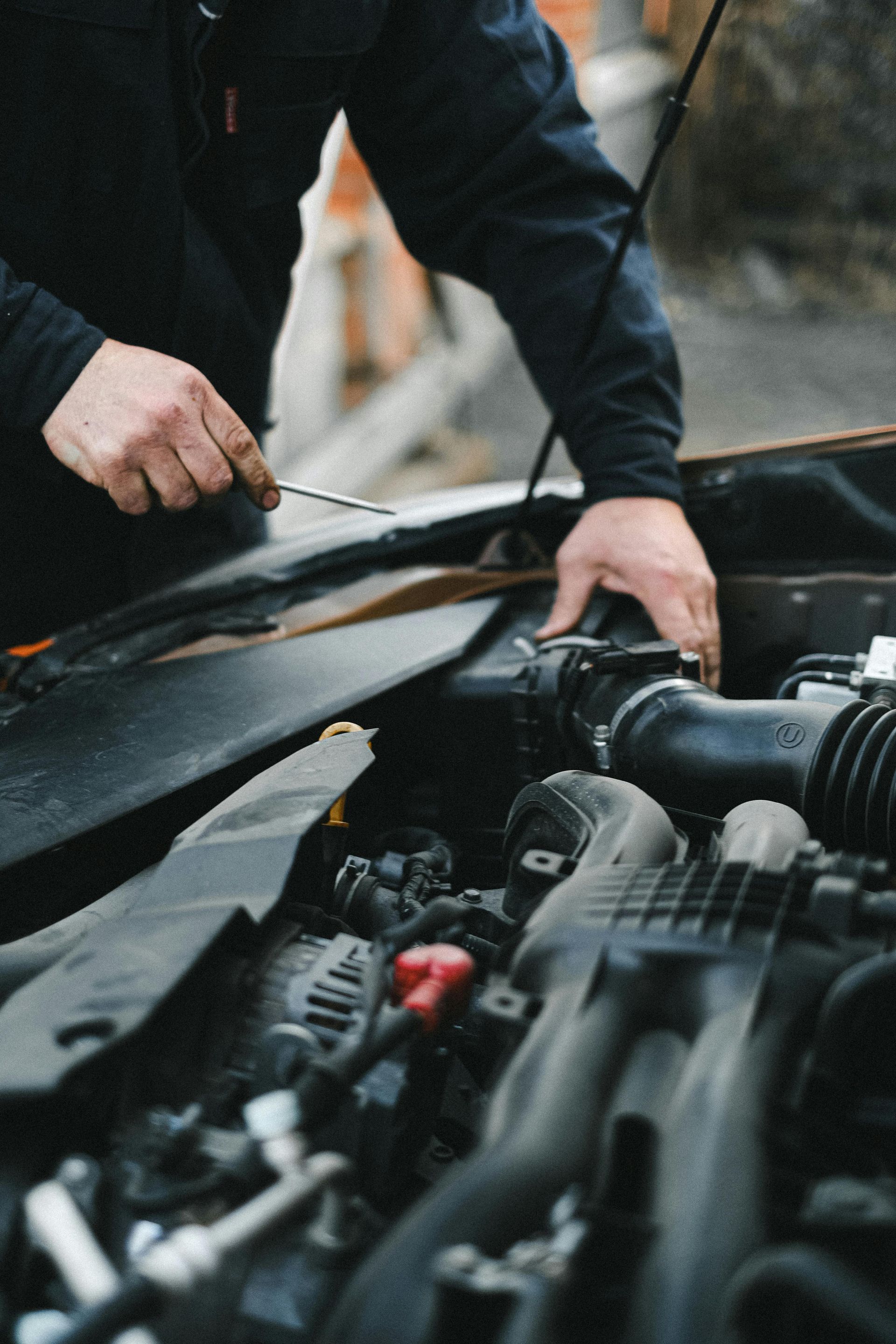 Person in dark clothing working on a car engine, holding a tool, outdoors.