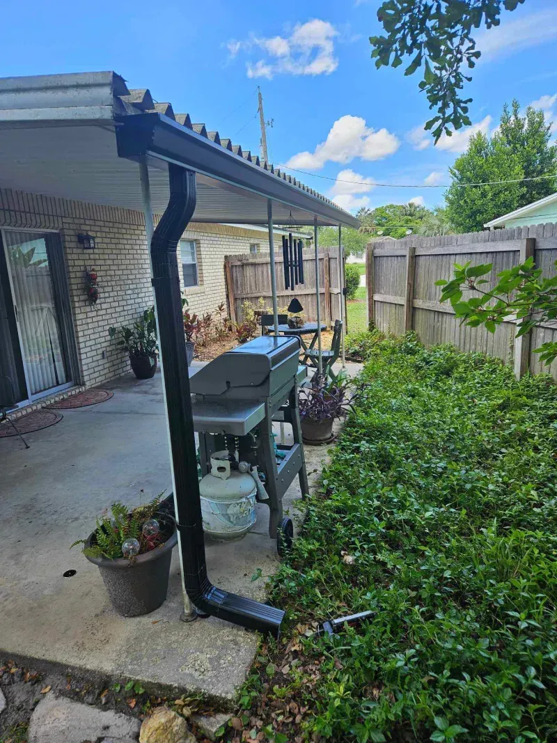 A covered patio with a grill and a drainpipe in the backyard of a house.