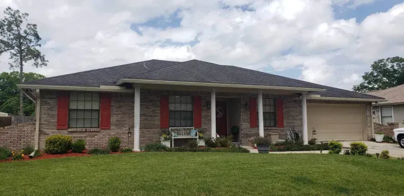 A brick house with a gray roof and red shutters is sitting on top of a lush green lawn.