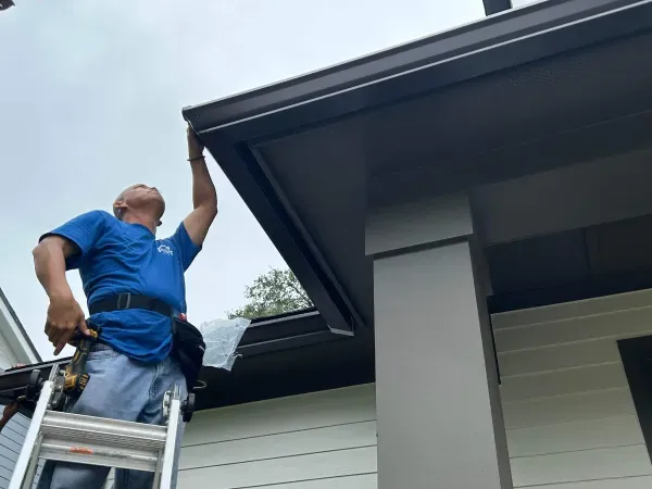 A man is standing on a ladder fixing a gutter on the side of a house.