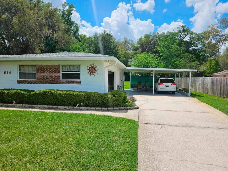 A white car is parked under a canopy in front of a white house.