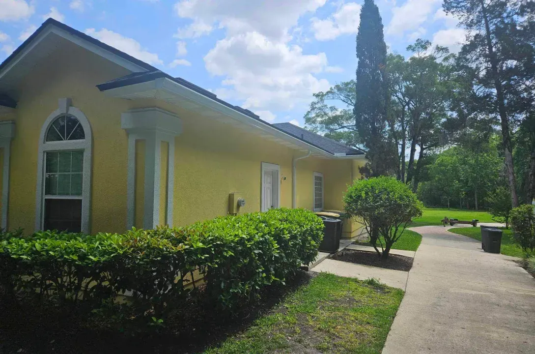 A yellow house with a walkway leading to it and trees in the background.