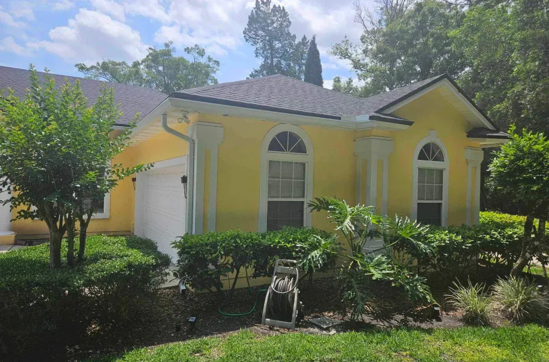 A yellow house with a gray roof is surrounded by trees and bushes.