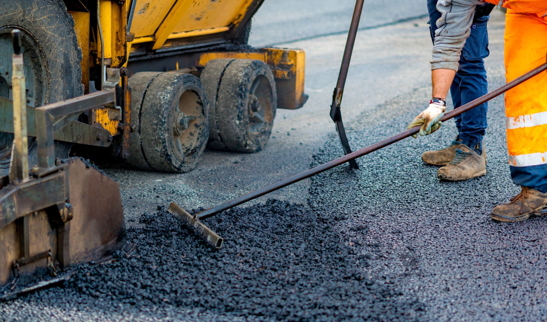 Construction worker raking fresh black asphalt beside a paving machine and dump truck
