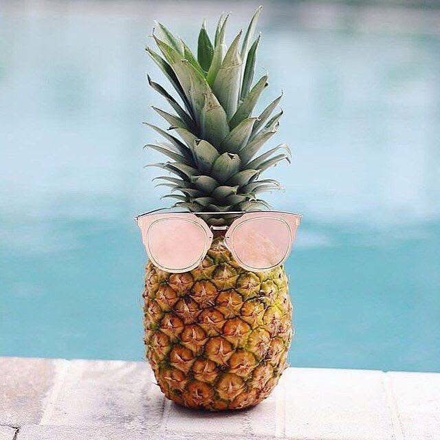 A whole pineapple wearing rose-gold sunglasses, sitting on the edge of a swimming pool.
