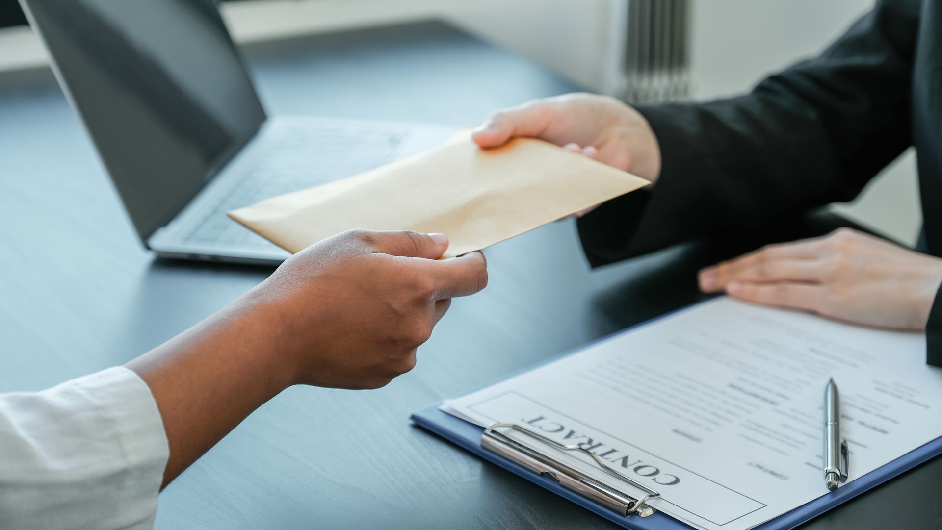 Hands exchanging an envelope over a contract on a desk, near a laptop.