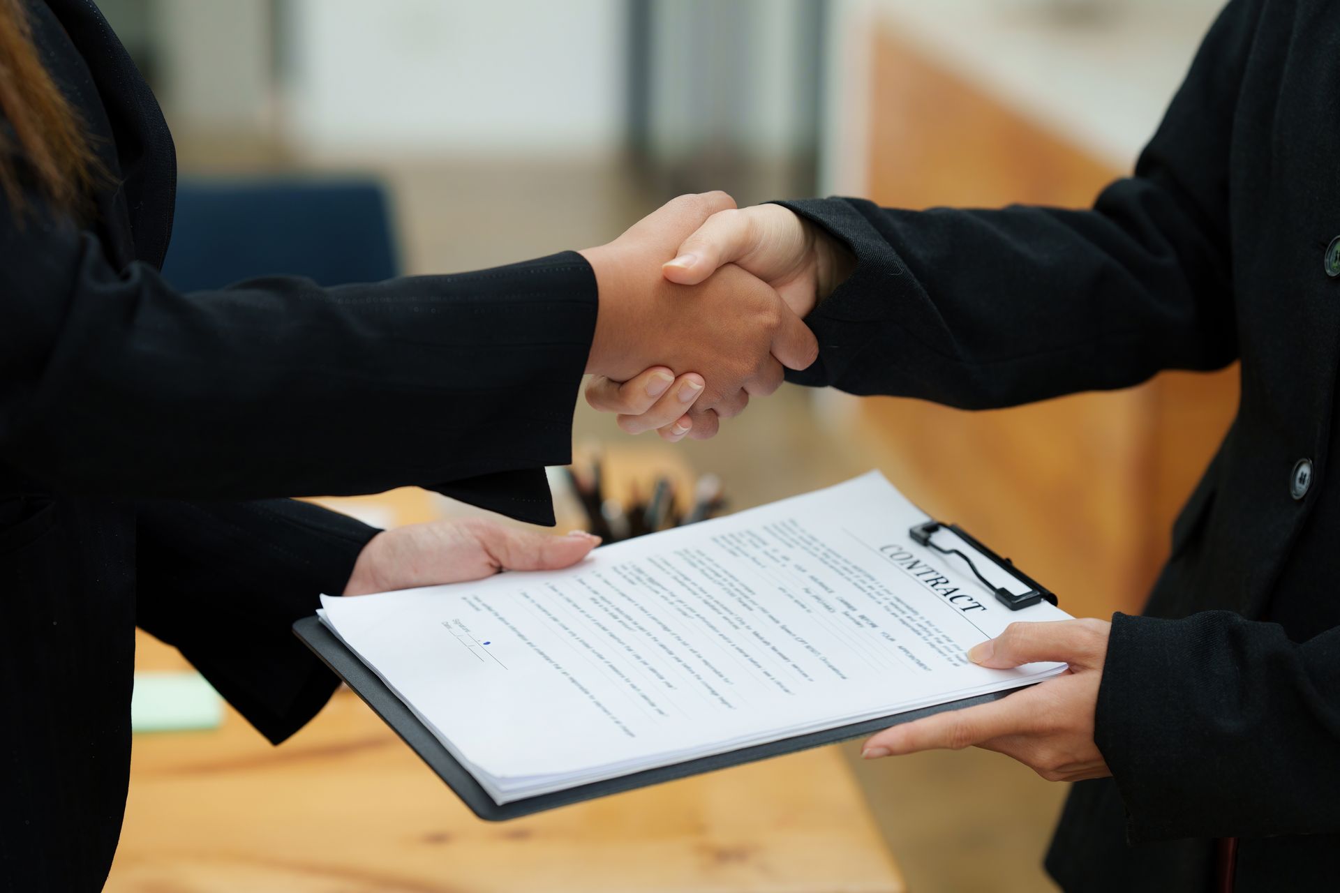 Two people shaking hands over a contract on a clipboard.