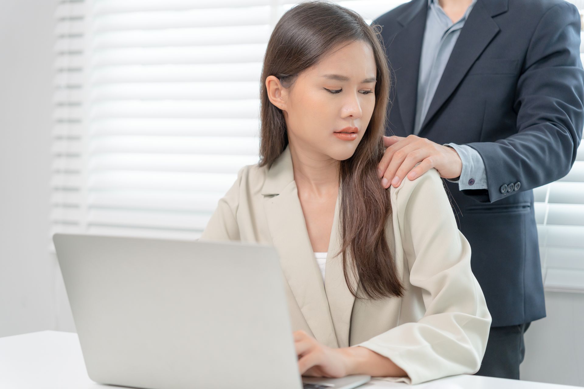 Man's hand on woman's shoulder; she looks uncomfortable while using a laptop. Office setting, window in background.